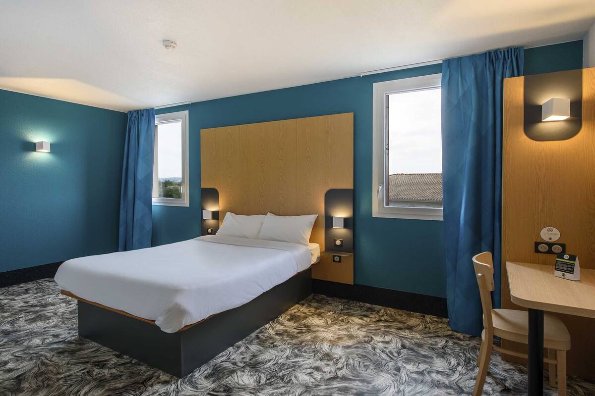 Ett hotellrum med en säng, ett skrivbord, en stol och ett fönster med gardiner.