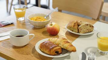 Une table en bois dressée pour le petit-déjeuner, avec des croissants, une pomme, du jus d’orange et des céréales.