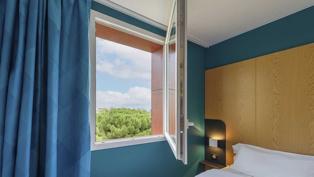 Una habitación de hotel con cama, mesita de noche, ventana con cortinas y vistas a zonas verdes.