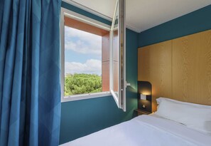 A hotel room with a bed, a nightstand, a window with curtains, and a view of greenery outside. - B&B HOTEL Toulouse Purpan Zénith (Toulouse)