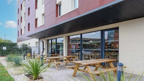 Un espacio al aire libre con mesas de madera y plantas en macetas, un edificio con grandes ventanales y suelo de grava.