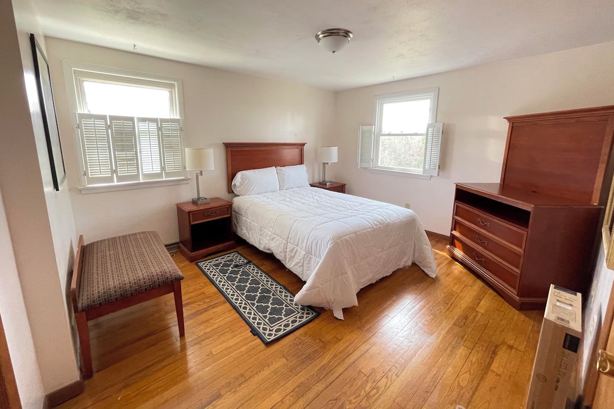 Quarto com cama, mesas de cabeceira, uma cadeira, cômoda e duas janelas com persianas.