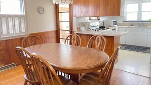 Un coin repas avec une table et des chaises en bois, un luminaire suspendu, et une cuisine dotée de meubles et d’appareils électroménagers blancs.