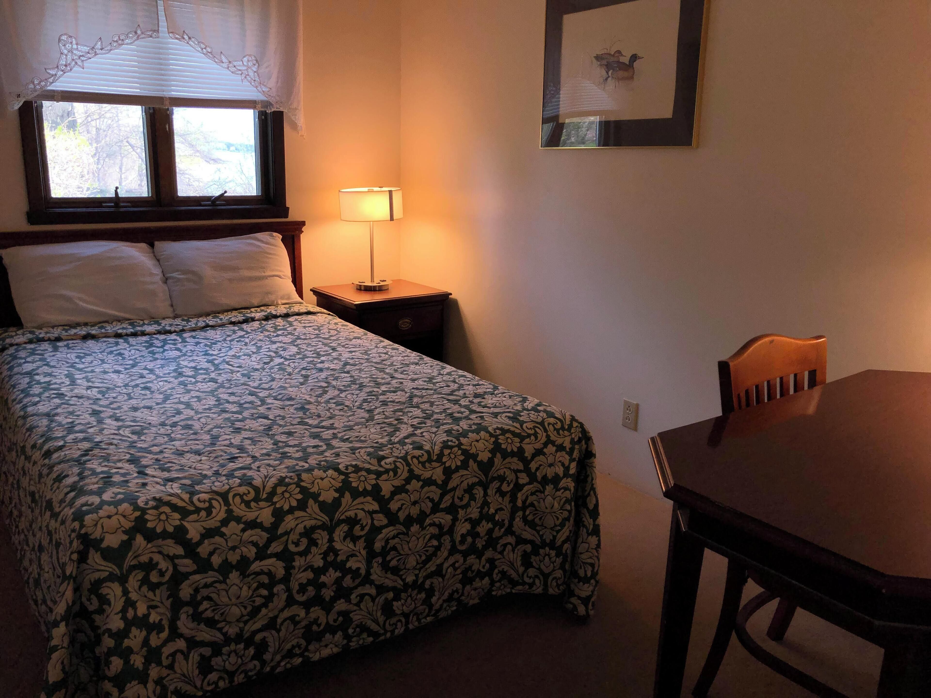 A neatly made bed with a patterned bedspread, a bedside table with a lamp, a framed picture on the wall, and a wooden table and chair in the corner.