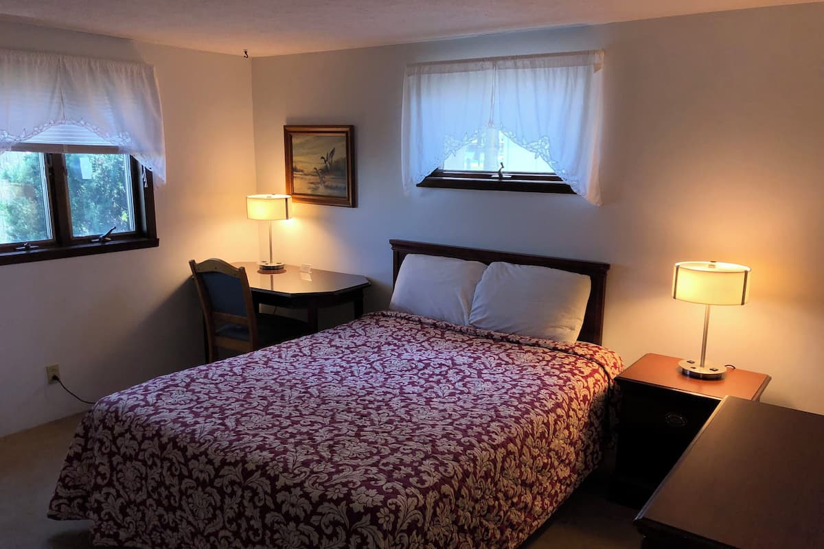 Quarto pequeno e bem arrumado com uma cama, duas luminárias de mesa, uma escrivaninha e uma cadeira.