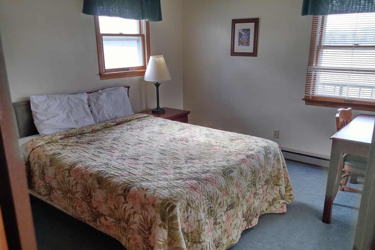 Quarto com cama, mesa de cabeceira, abajur, cadeira e janela com persianas.