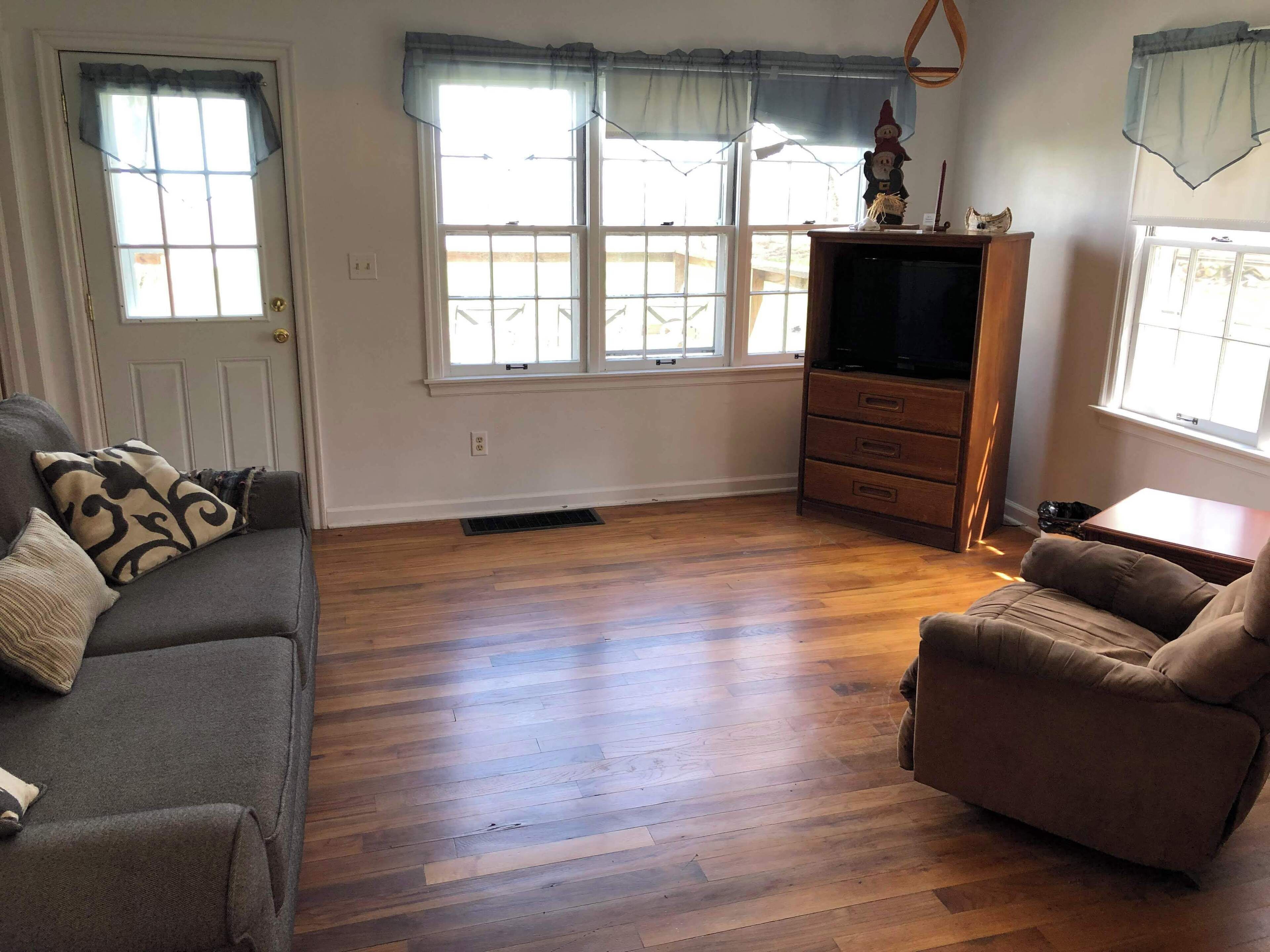 A living room with hardwood flooring, a grey sofa, a brown armchair, a wooden TV stand, and a window with curtains.