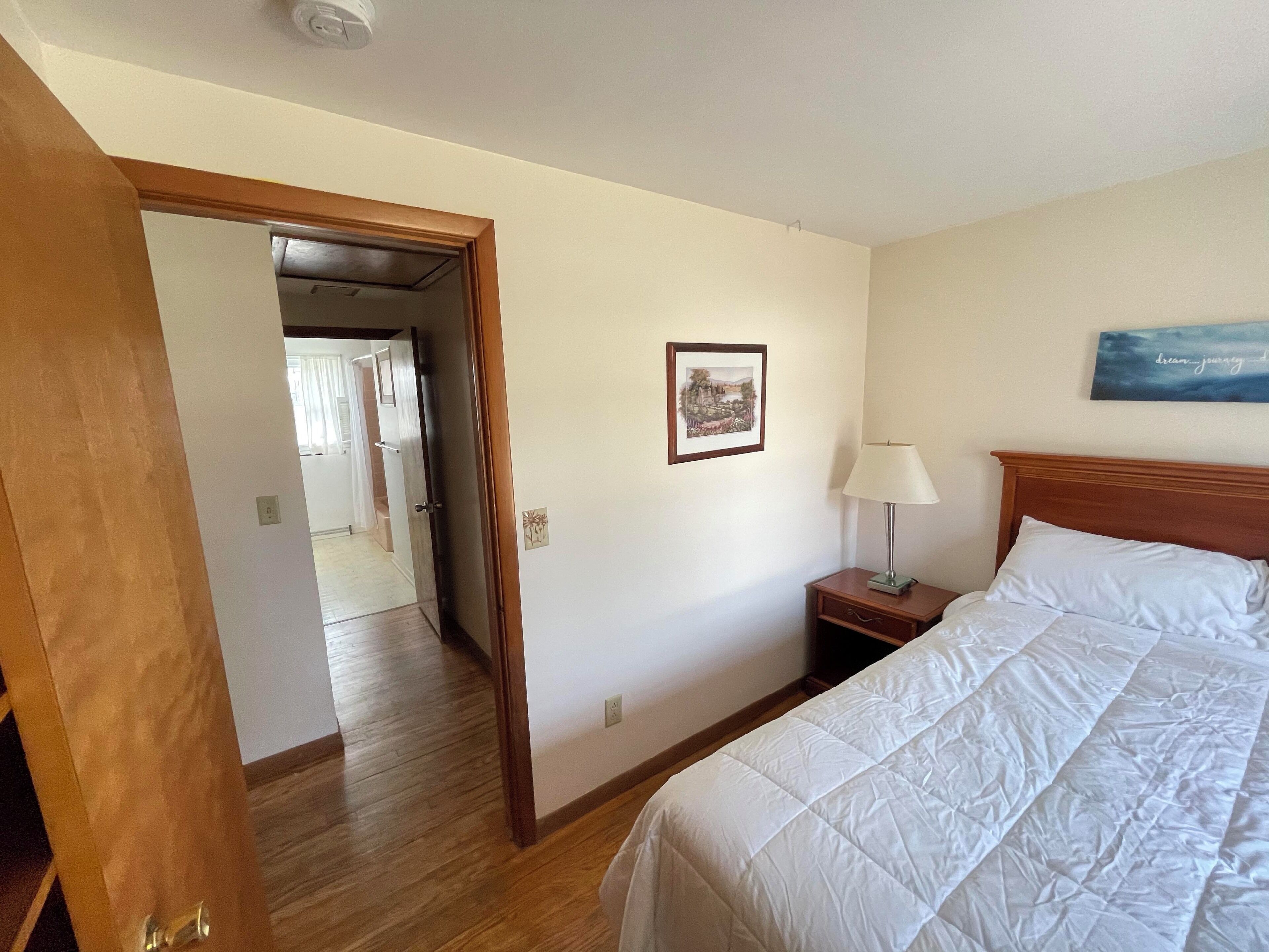 A hotel room with a bed, a nightstand with a lamp, a framed picture on the wall, and a wooden door leading to another room.