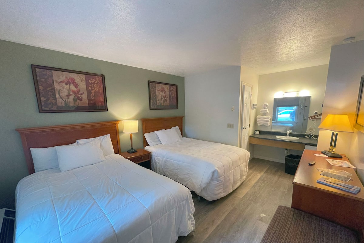 Quarto de hotel com duas camas, uma escrivaninha, um espelho e um banheiro visível através de uma porta aberta.