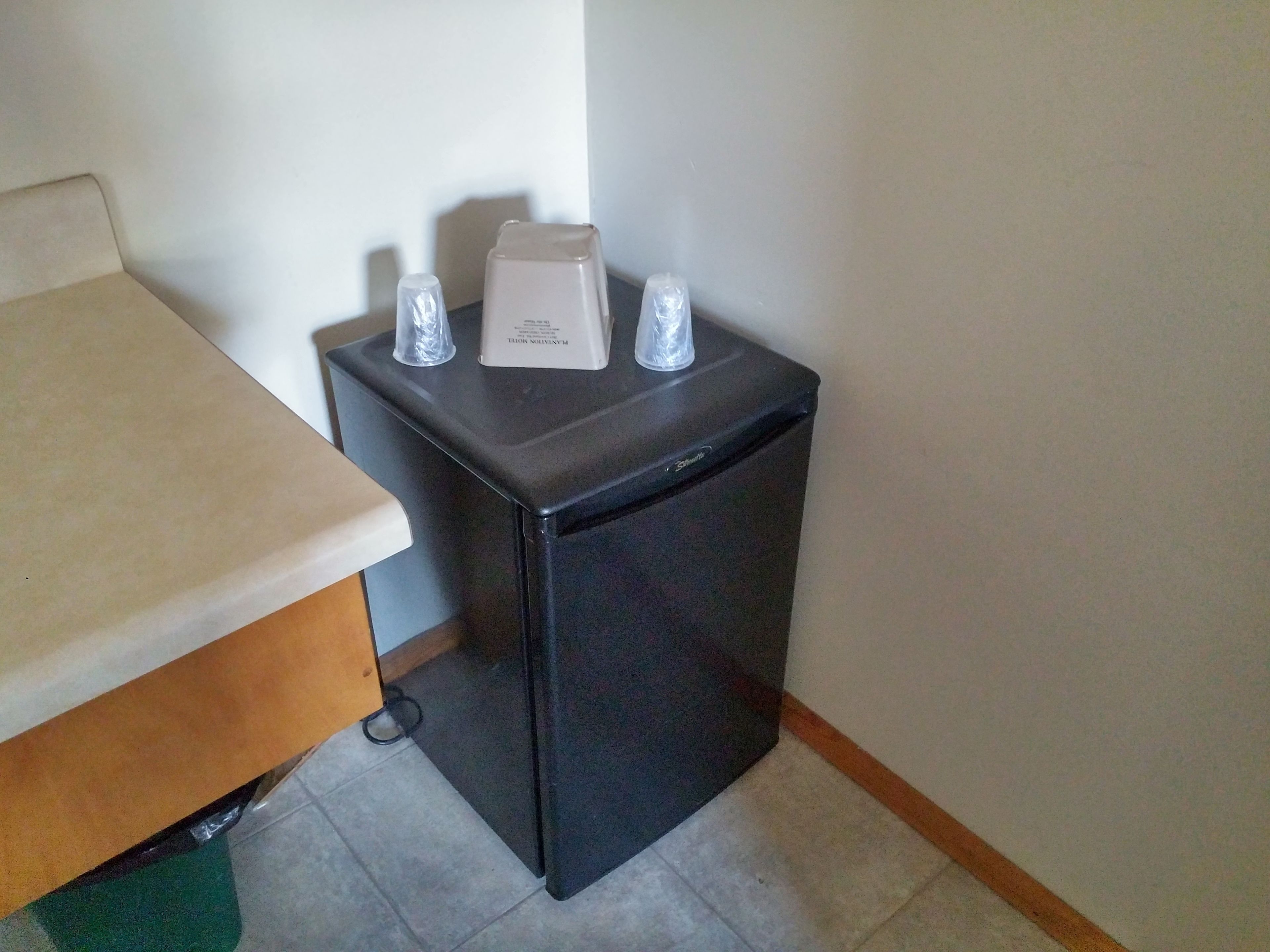 Un refrigerador compacto negro con un panel de control blanco en la parte superior, ubicado contra una pared en una esquina.