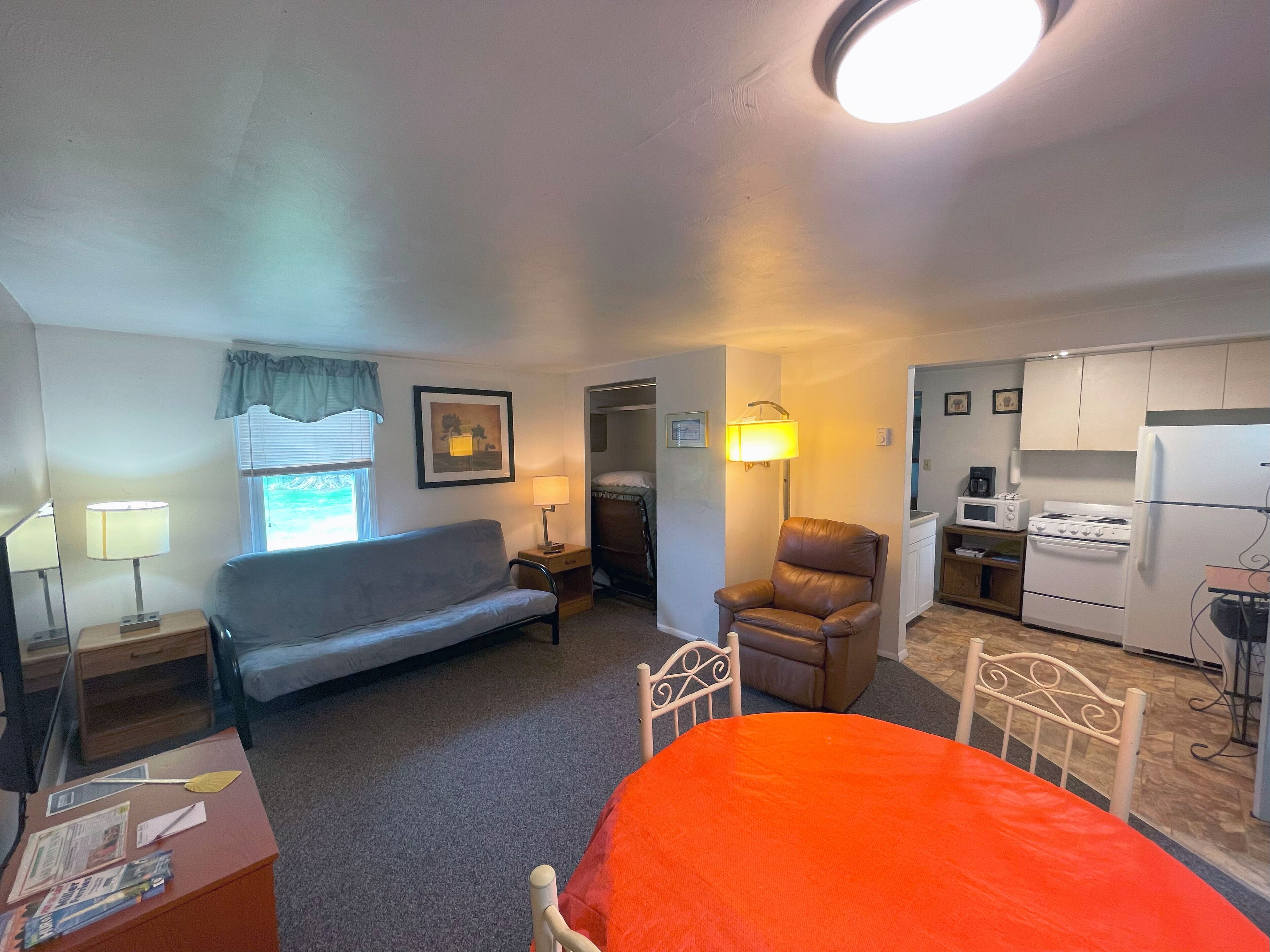 Habitación de hotel compacta con cama, sofá, silla, escritorio, lámpara, un cuadro en la pared, ventana con cortinas y kitchenette con estufa y microondas.