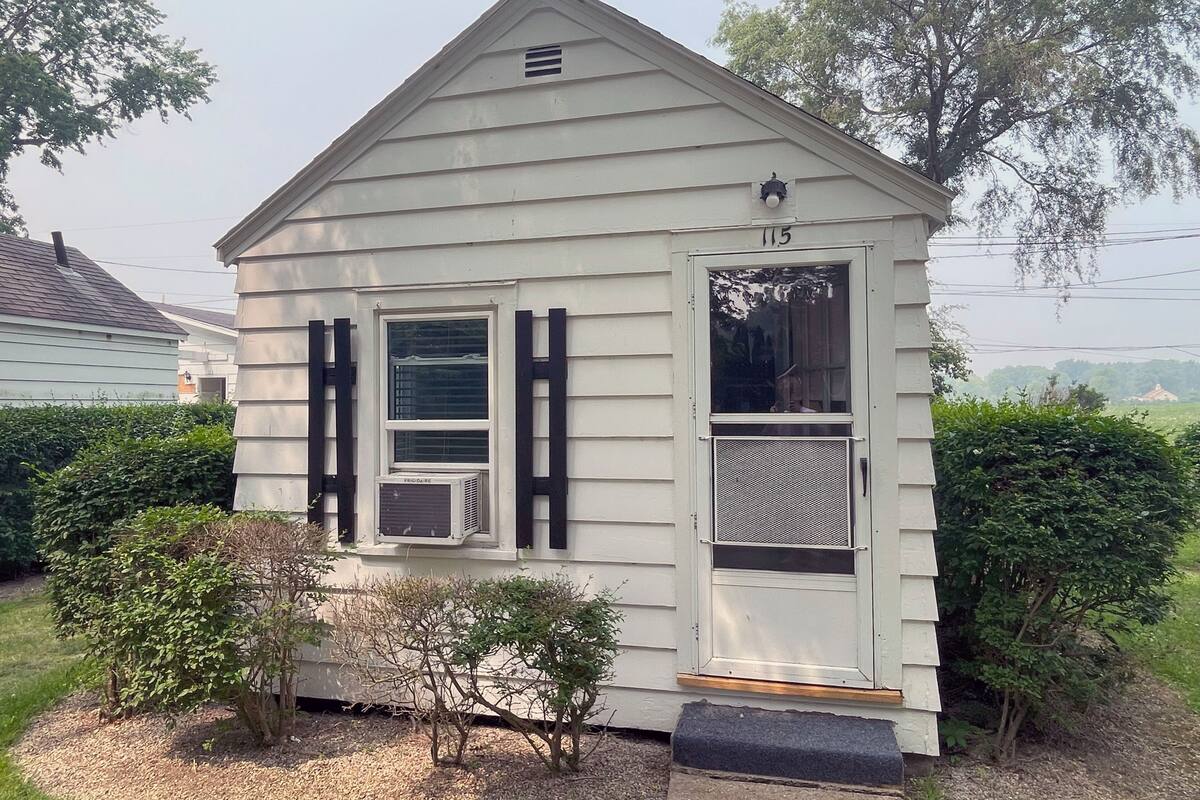 Uma casa pequena, térrea, com telhado em duas águas, varanda frontal e o número '115' na porta.