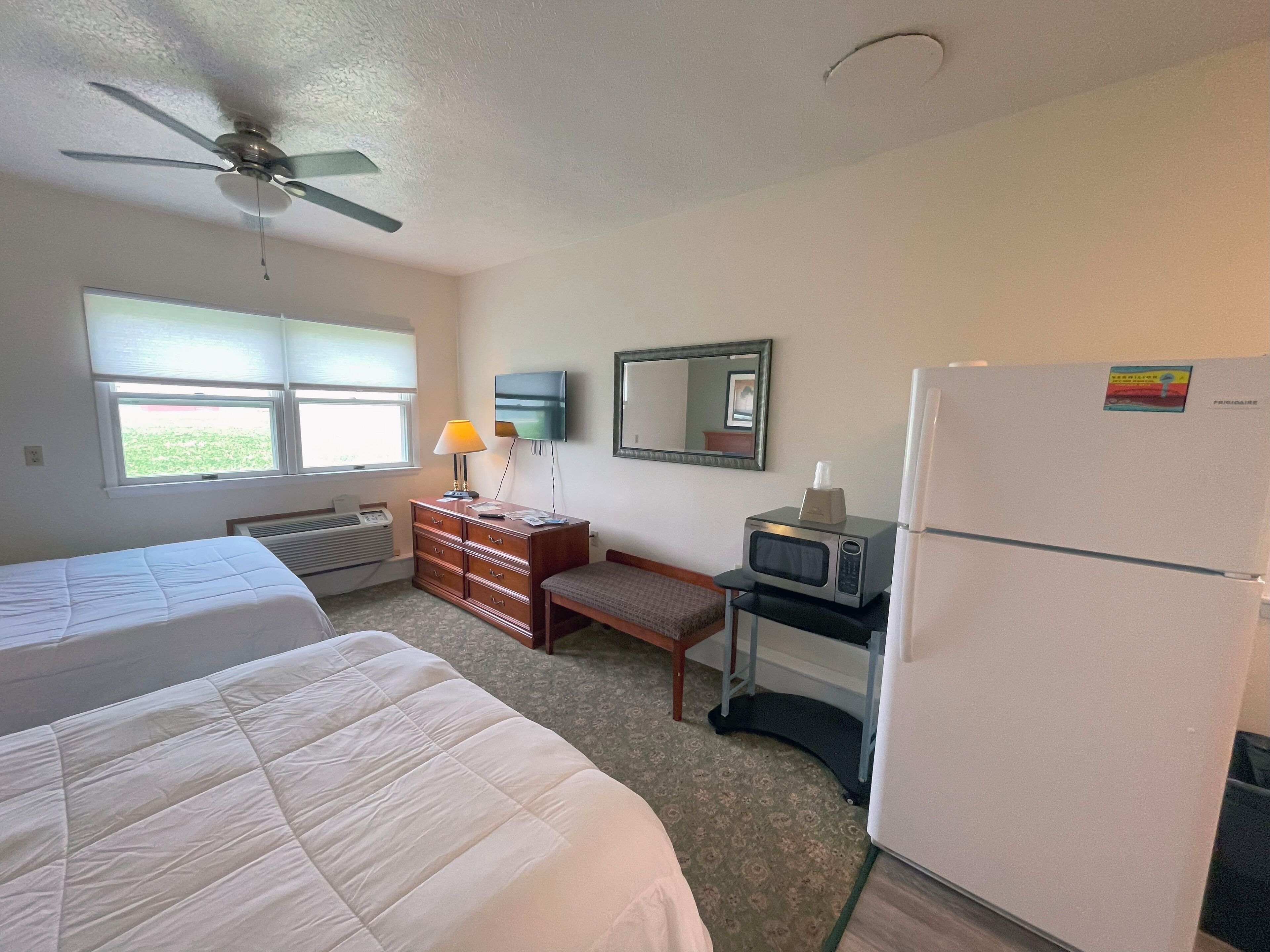 A hotel room with two beds, a ceiling fan, a desk with a microwave, a refrigerator, and a window with blinds.
