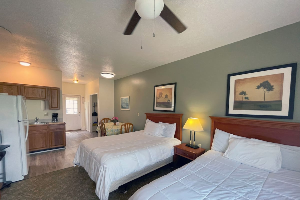 Quarto de hotel com duas camas, área de refeições, cozinha compacta e ventilador de teto.
