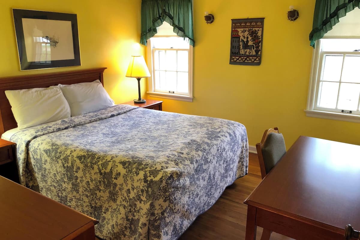 Quarto de hotel com cama, escrivaninha, luminária, janela com cortinas e detalhes decorativos nas paredes.