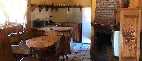 A rustic kitchen with a wooden table and chairs, a brick fireplace, and a hanging light fixture.