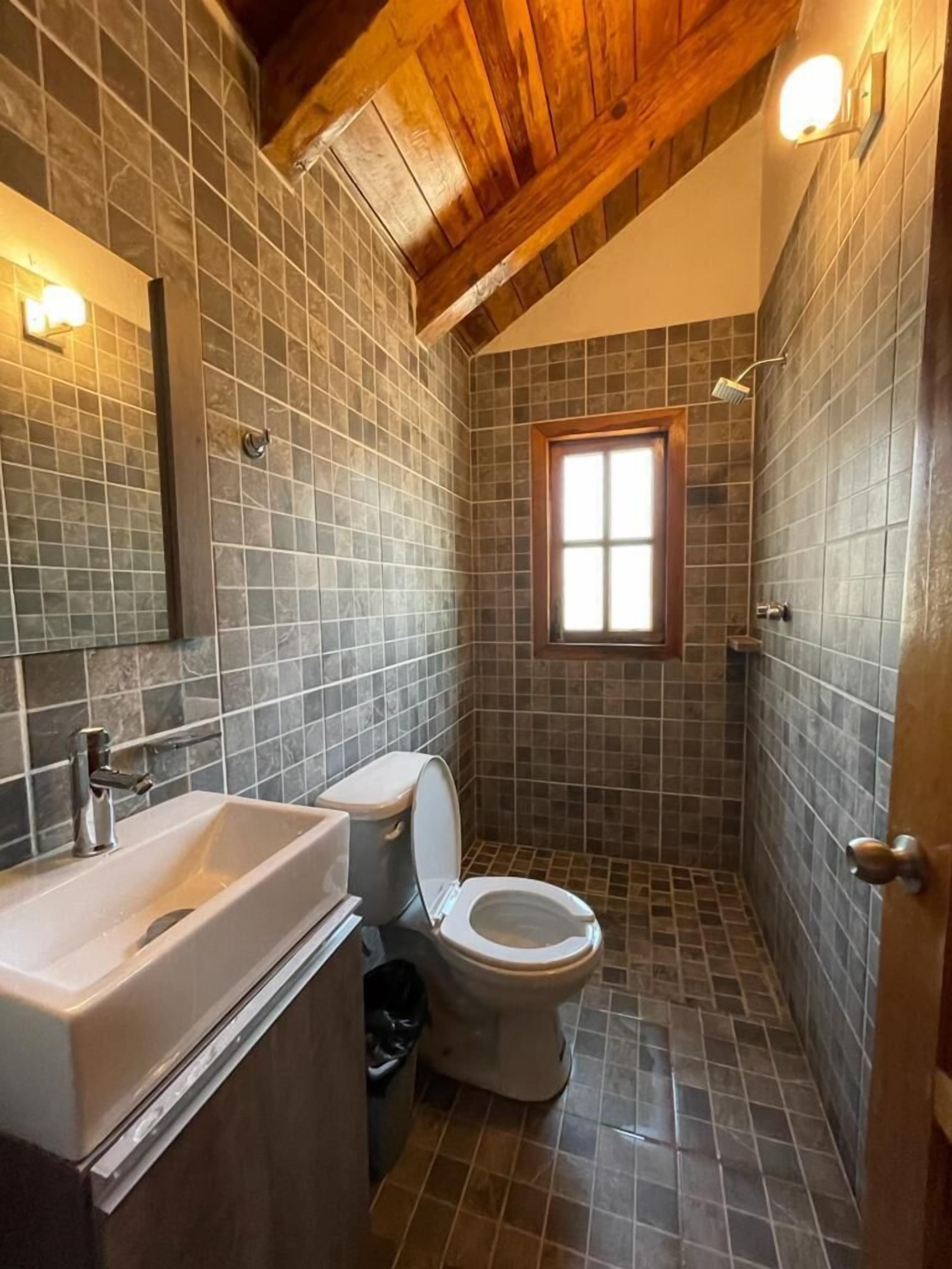 Un bagno compatto con soffitto in legno, pareti piastrellate, una finestra, un lavandino e un water.