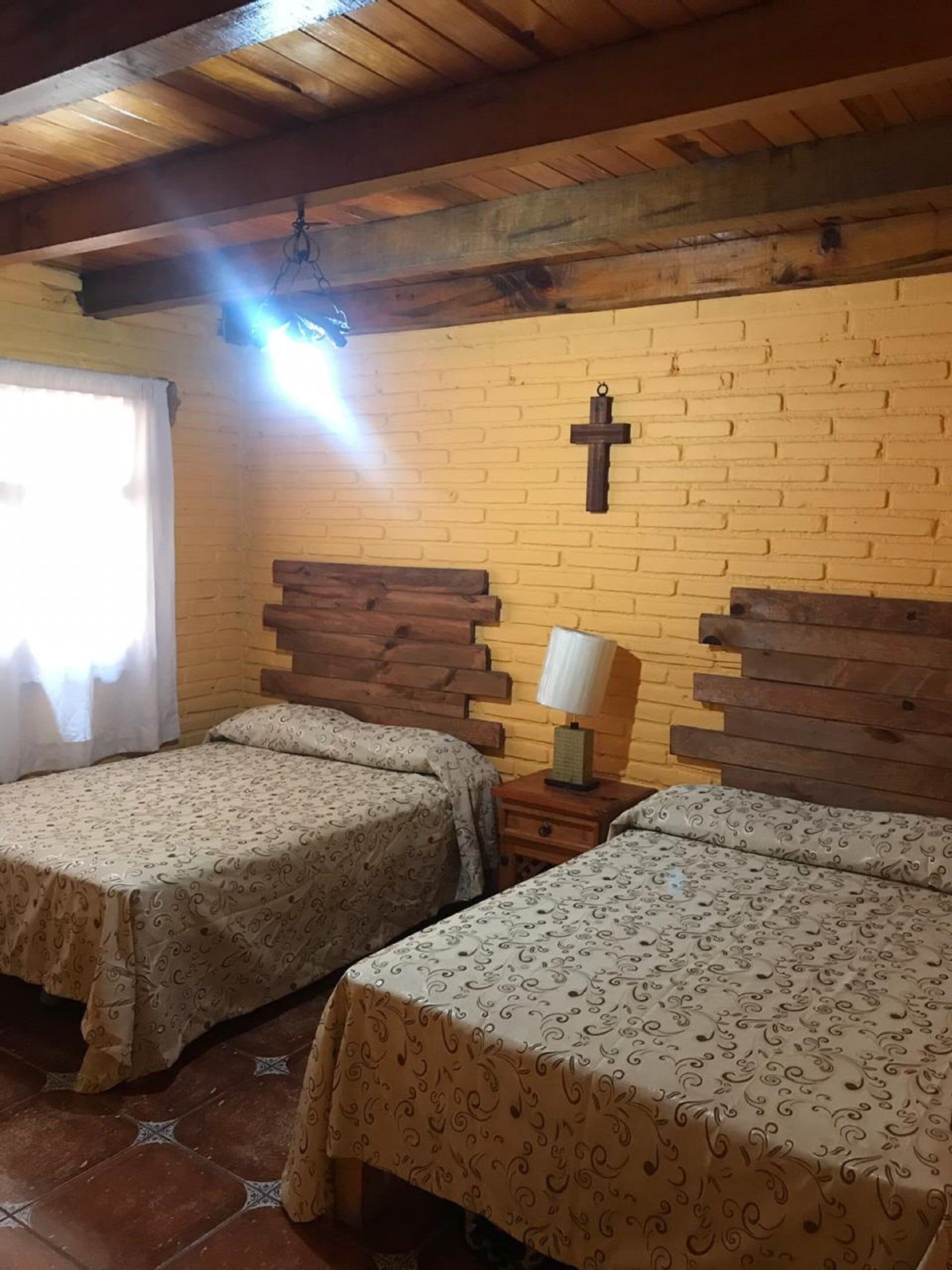 Ein Zimmer mit zwei Betten, Holz-Kopfteilen, einem Kruzifix an der Wand, einer Lampe und einem Fenster mit Vorhängen.