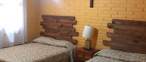 A room with two beds, wooden headboards, a cross on the wall, a lamp, and a window with curtains.