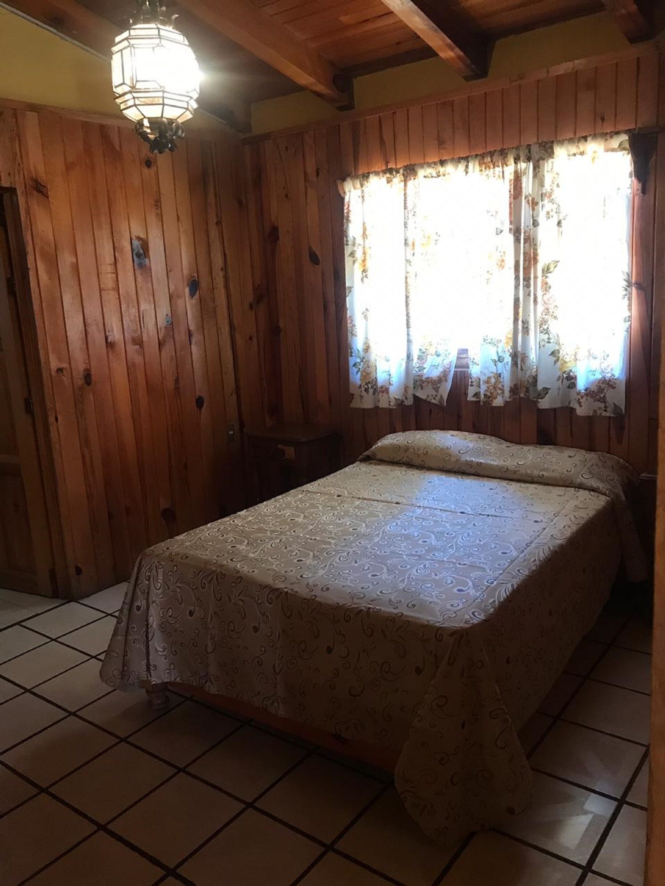 Une pièce en bois avec un lit, une fenêtre ornée de rideaux à motifs floraux et un luminaire suspendu.