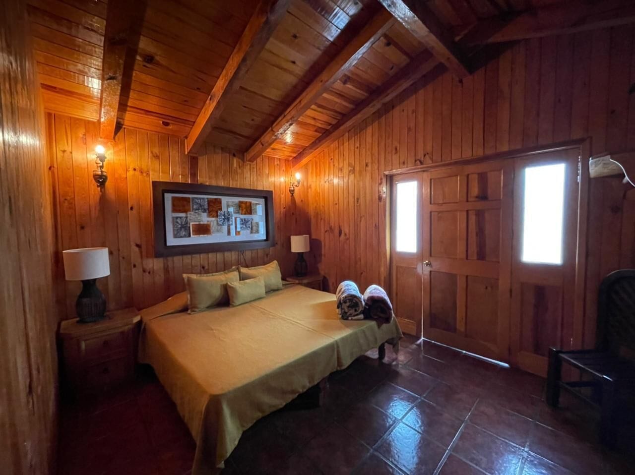 A wooden cabin bedroom with a bed, two nightstands, a wall-mounted picture, and a door with a window.