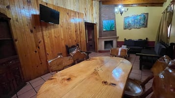 A wooden-paneled room with a large wooden table and chairs, a television mounted on the wall, and a fireplace.