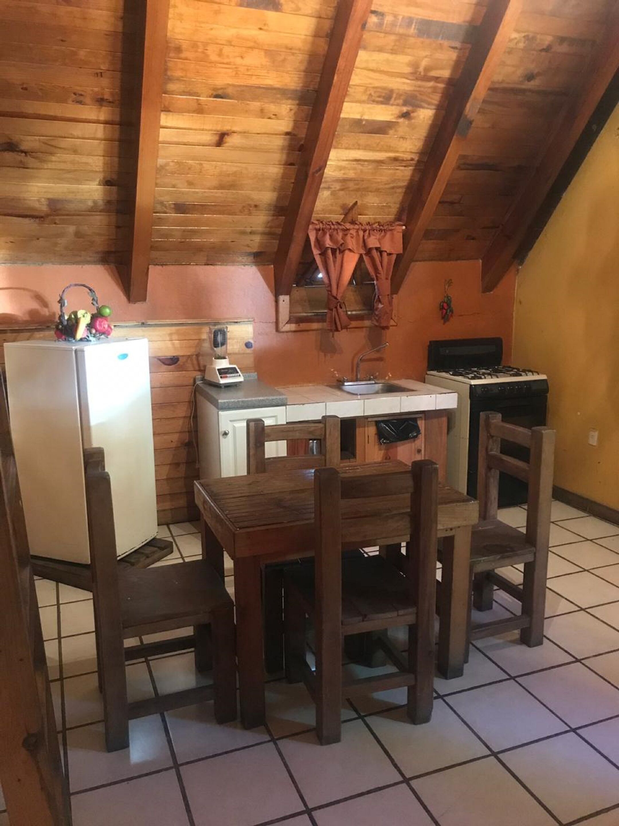 A small kitchen with a wooden table and chairs, a refrigerator, a stove, and a window with curtains.
