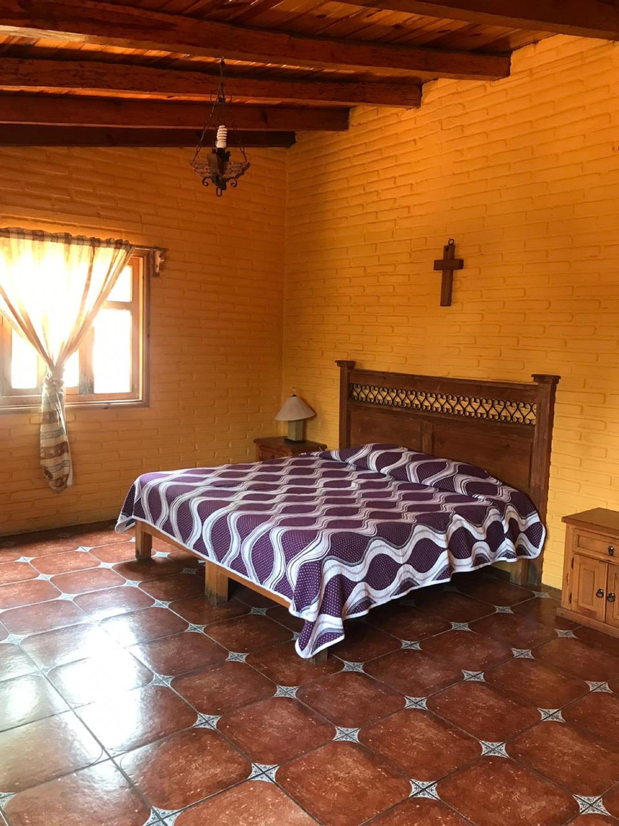 A bedroom with a wooden bed, a brick wall, a cross, and a window with curtains.