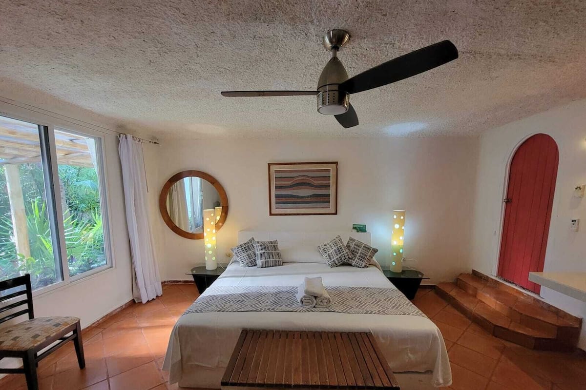 A bedroom with a bed, a ceiling fan, a bedside table with a lamp, a chair, a window with curtains, and a red door.