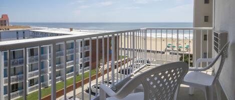 A balcony with white chairs, a view of the beach, and a building with multiple windows.