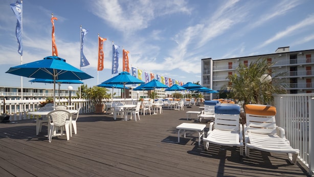 A wooden deck with white lounge chairs and blue umbrellas, flags, and a building in the background. - The Spinnaker Hotel (Ocean City)