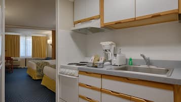 A compact hotel room with a kitchenette, a bed, a desk, and a view of the hotel lobby.