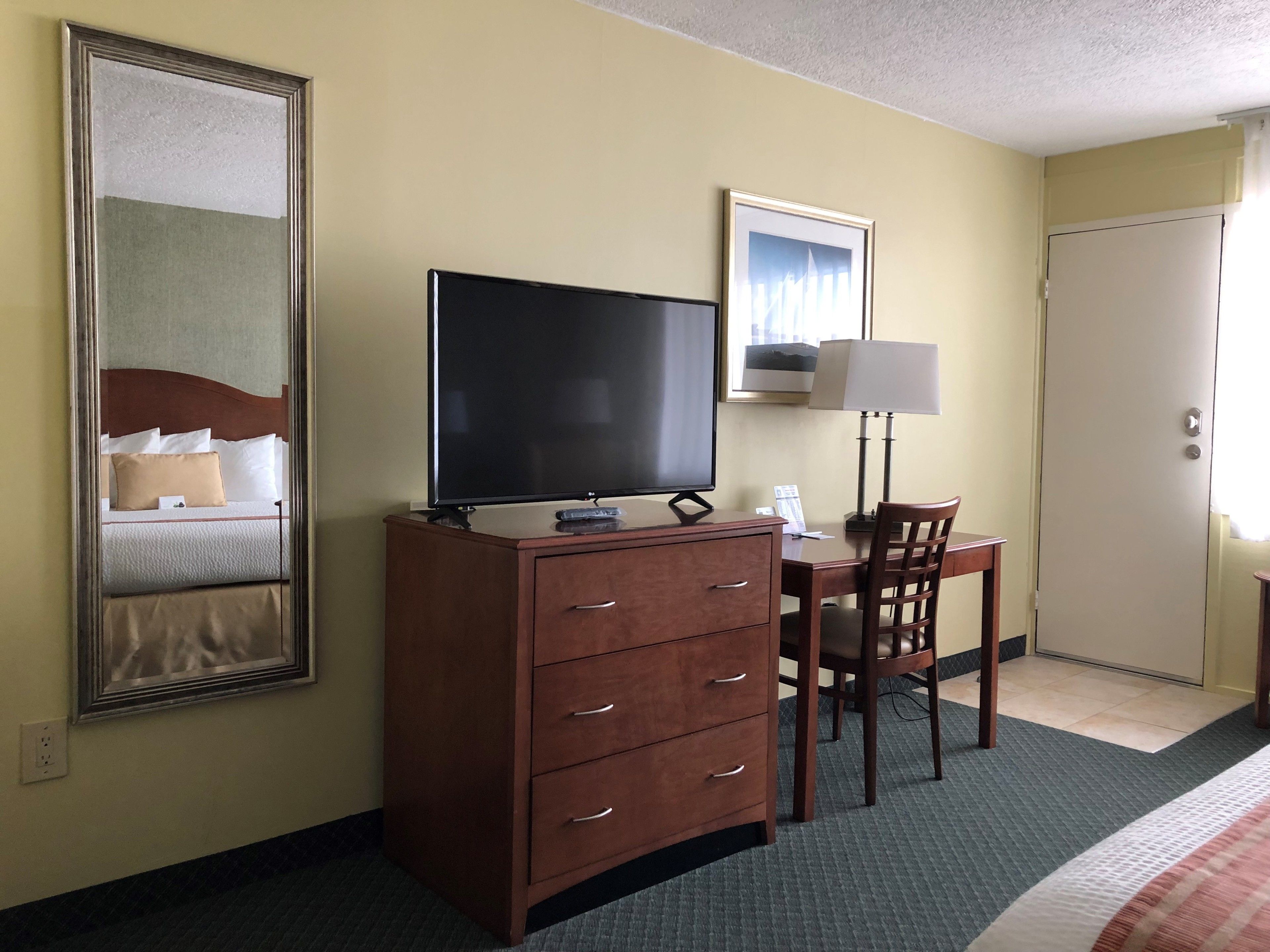 Une chambre d’hôtel avec un grand miroir, un téléviseur à écran plat posé sur une commode en bois, un bureau avec une chaise, une lampe et une porte.