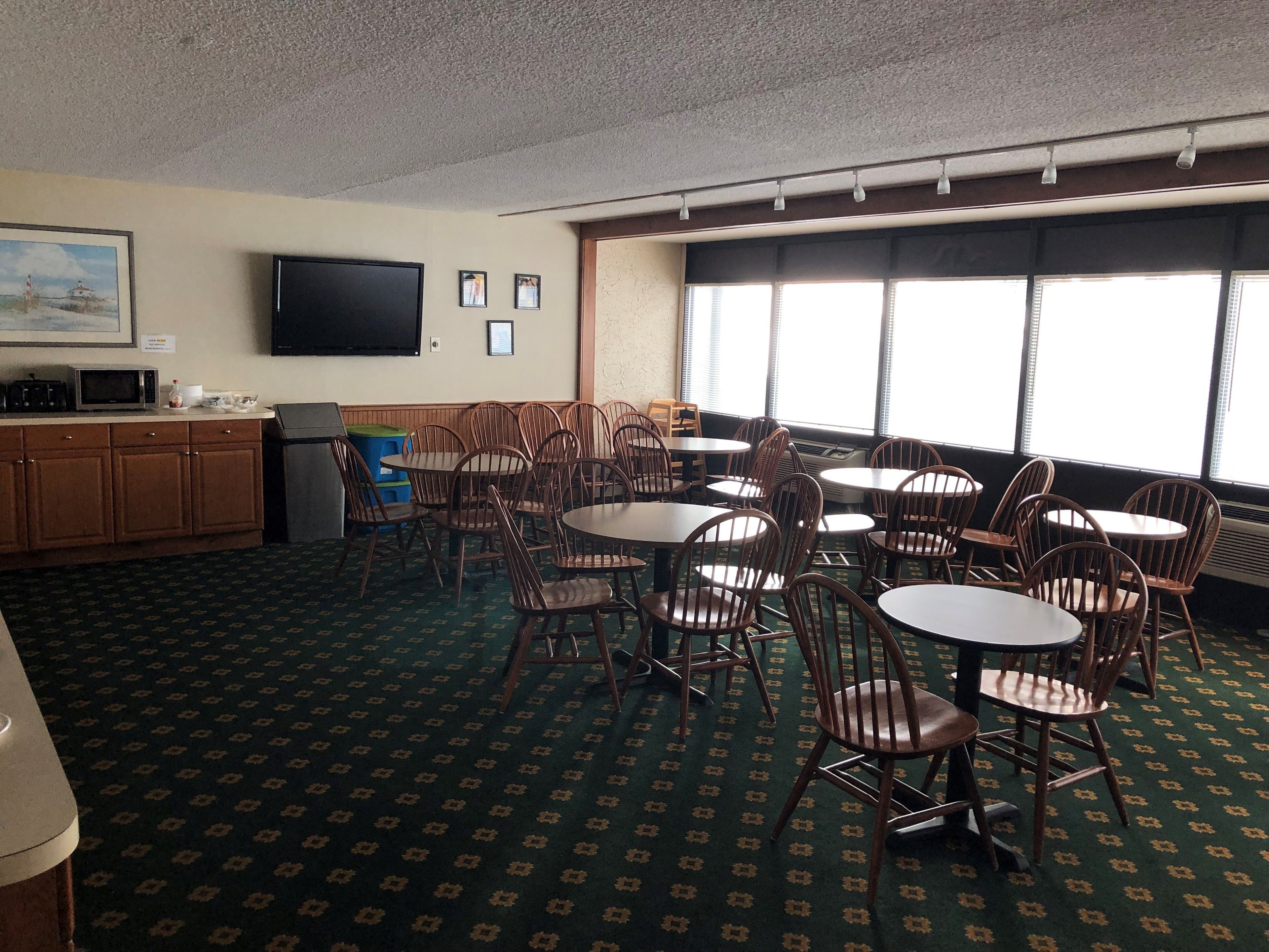 A hotel room with a dining area featuring round tables and wooden chairs, a kitchenette with a microwave, and a television mounted on the wall.