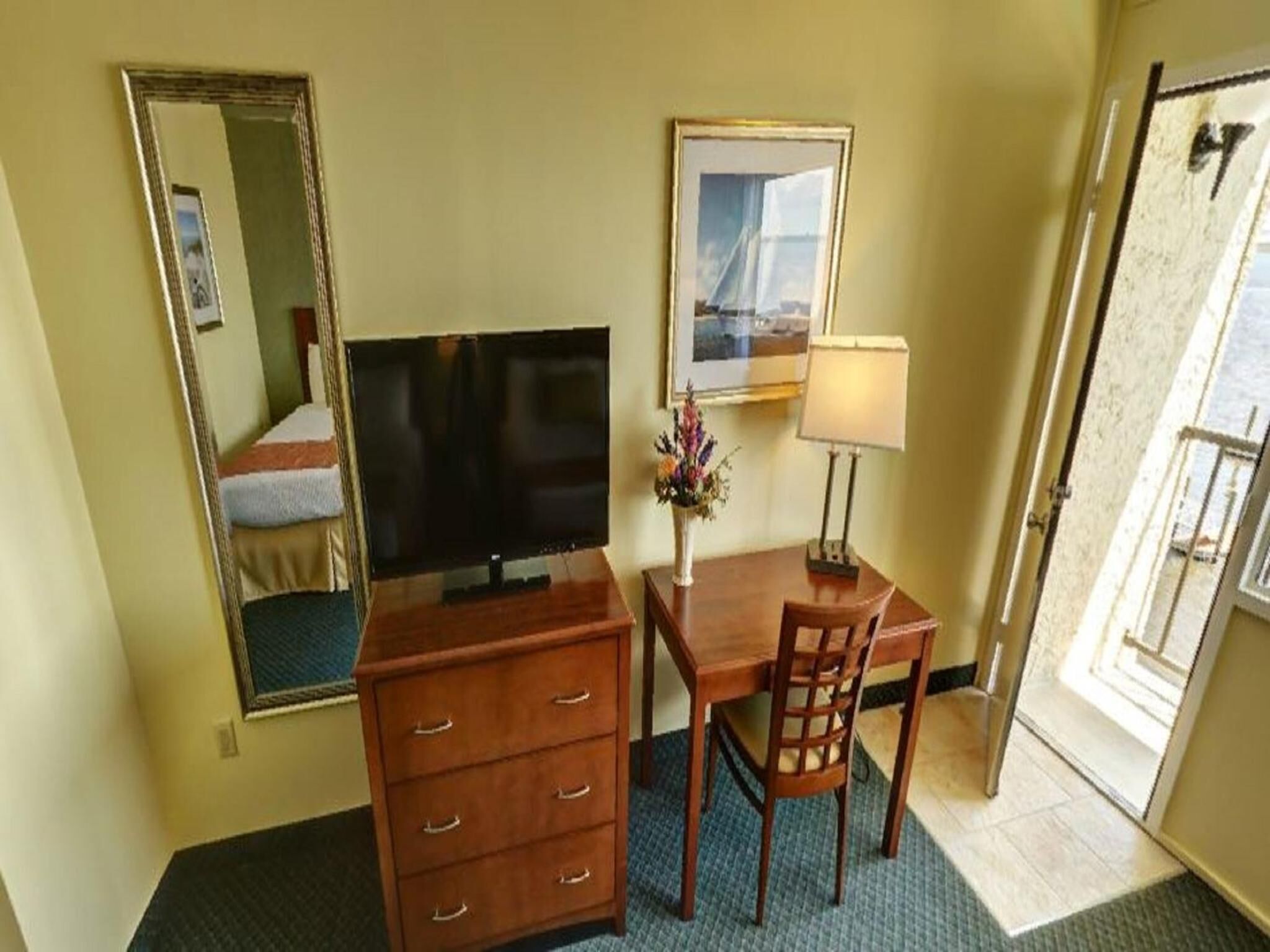 Una habitación de hotel con cama, televisión, escritorio con silla, cómoda, lámpara y un cuadro en la pared.