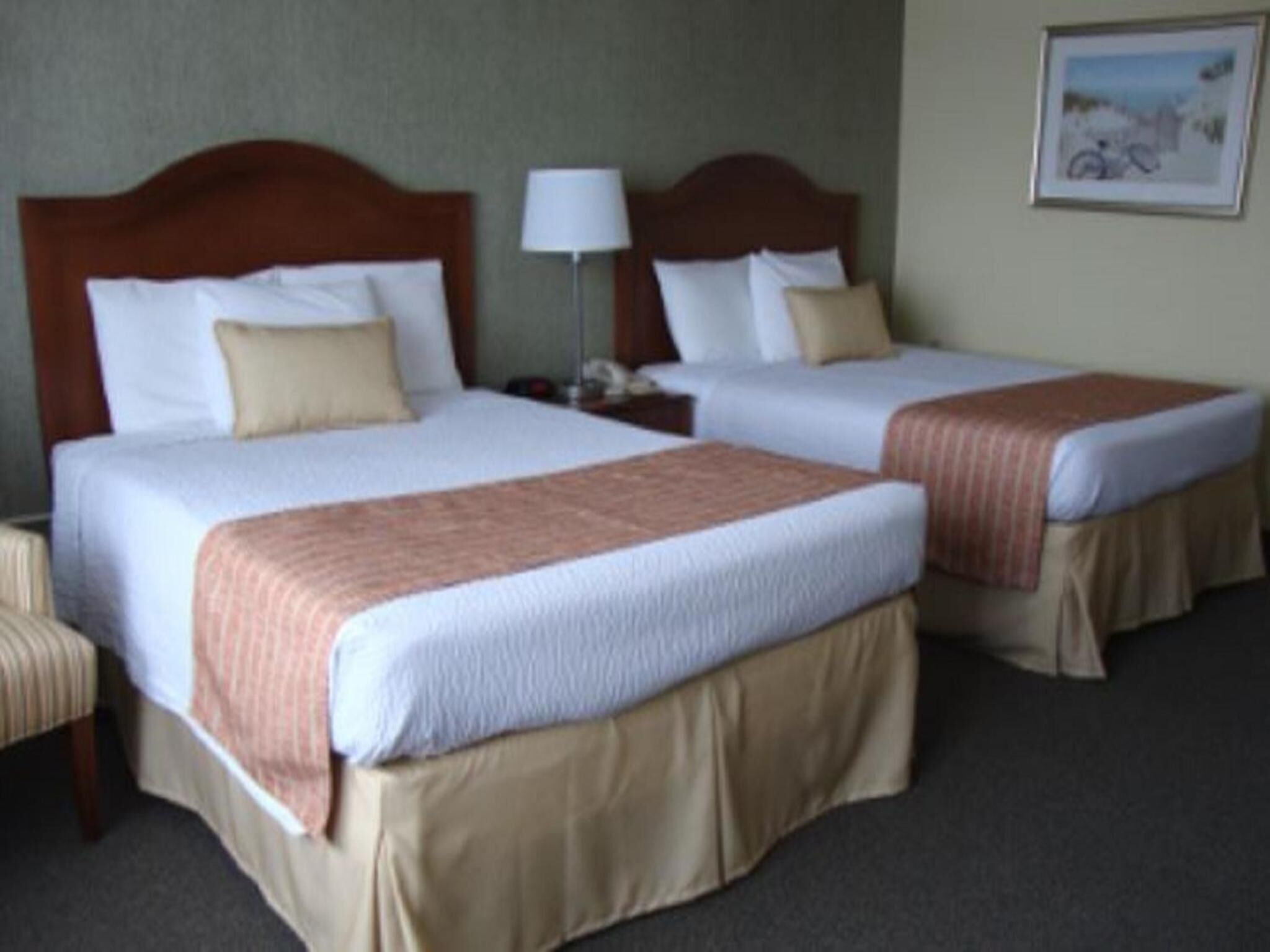 A hotel room with two beds, each with a wooden headboard, white linens, and a beige skirt. There is a nightstand with a lamp between the beds, and a framed picture on the wall.