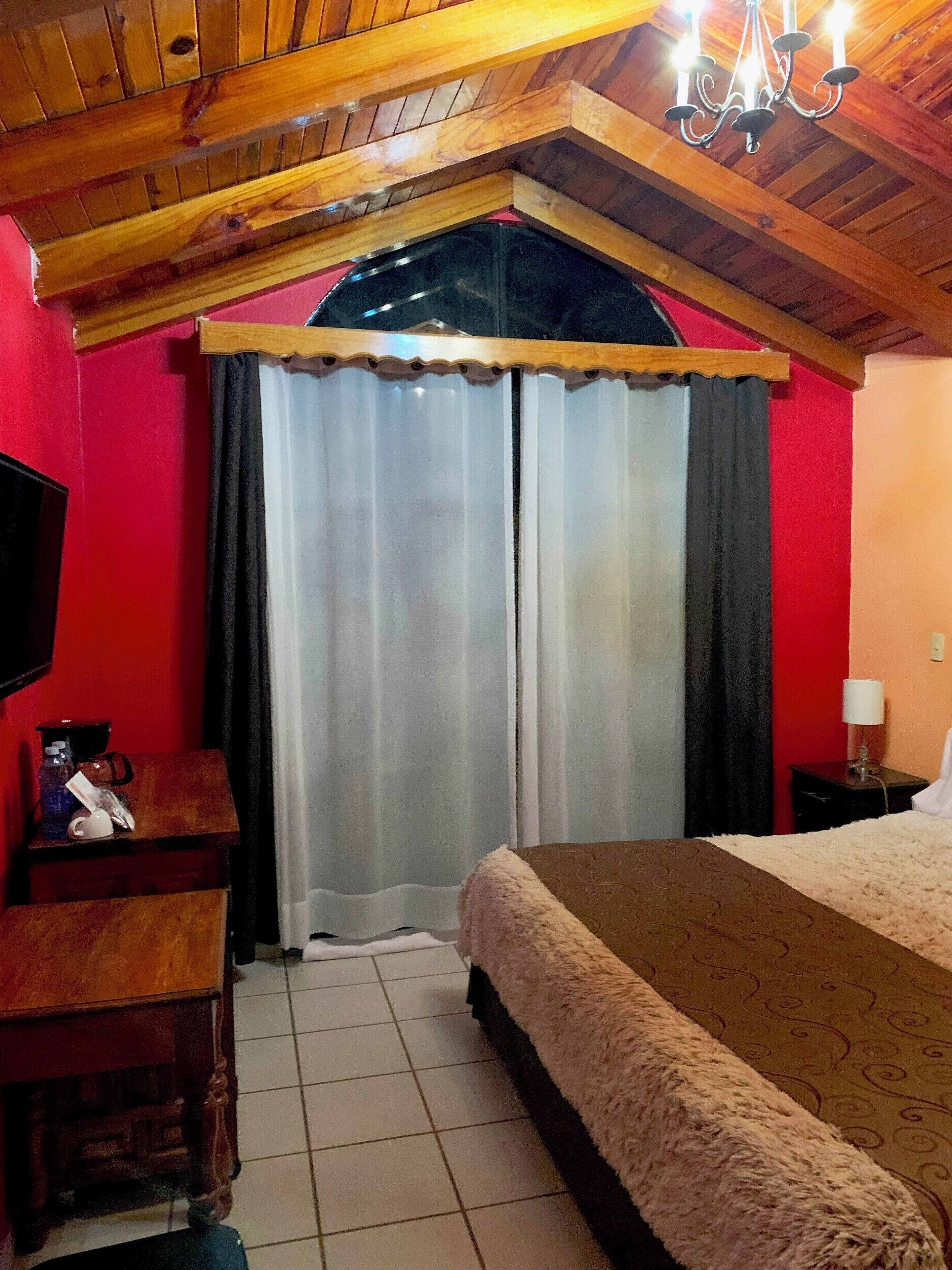 Habitación de hotel con techo de madera, una cama con manta mullida, una mesita de noche de madera con lámpara y una pared roja.