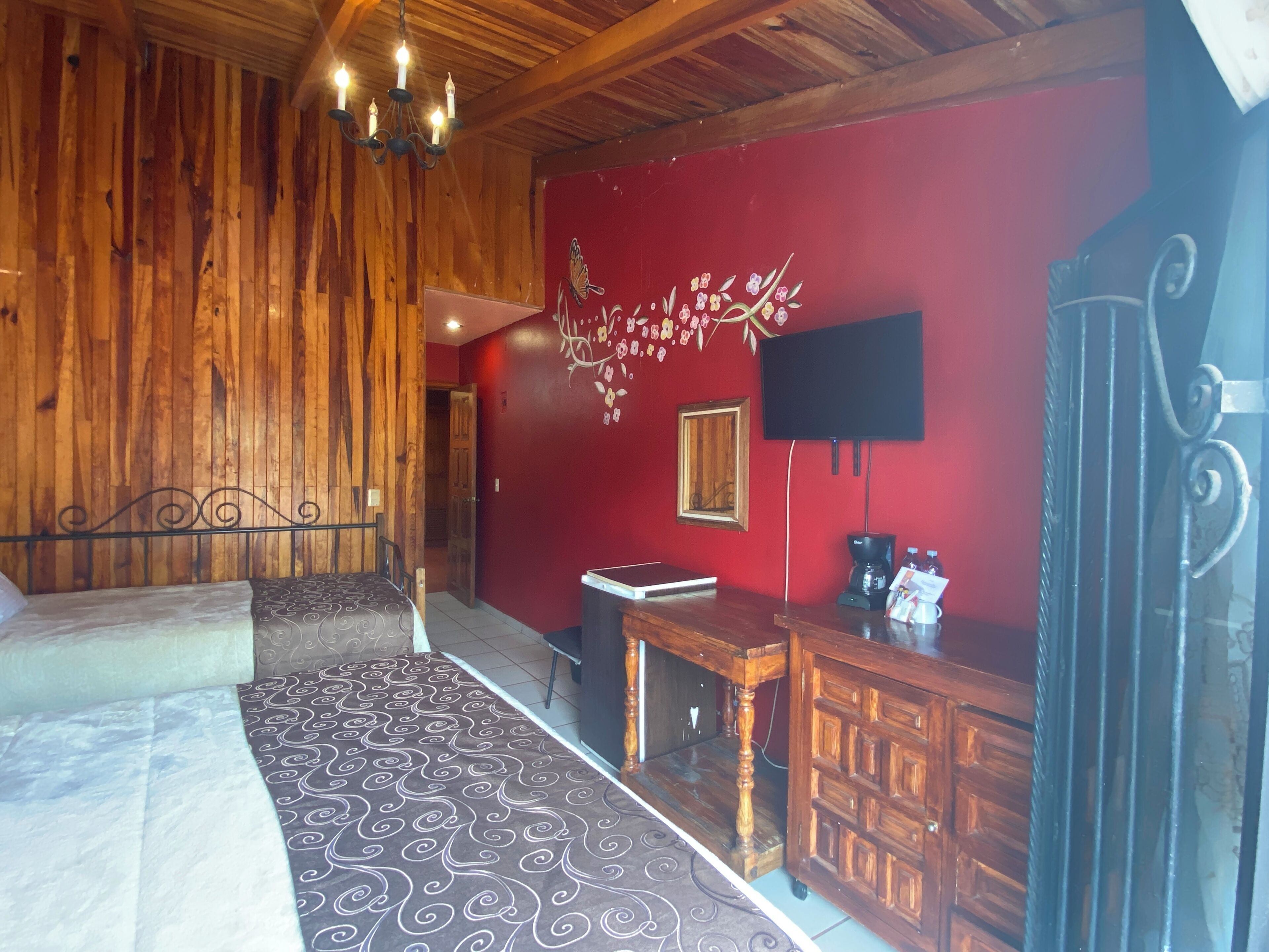 Ein Zimmer mit Holzboden, einem Bett mit gemusterten Textilien, einer roten Wand mit floralem Dekor, einem Fernseher und einer Kaffeemaschine.