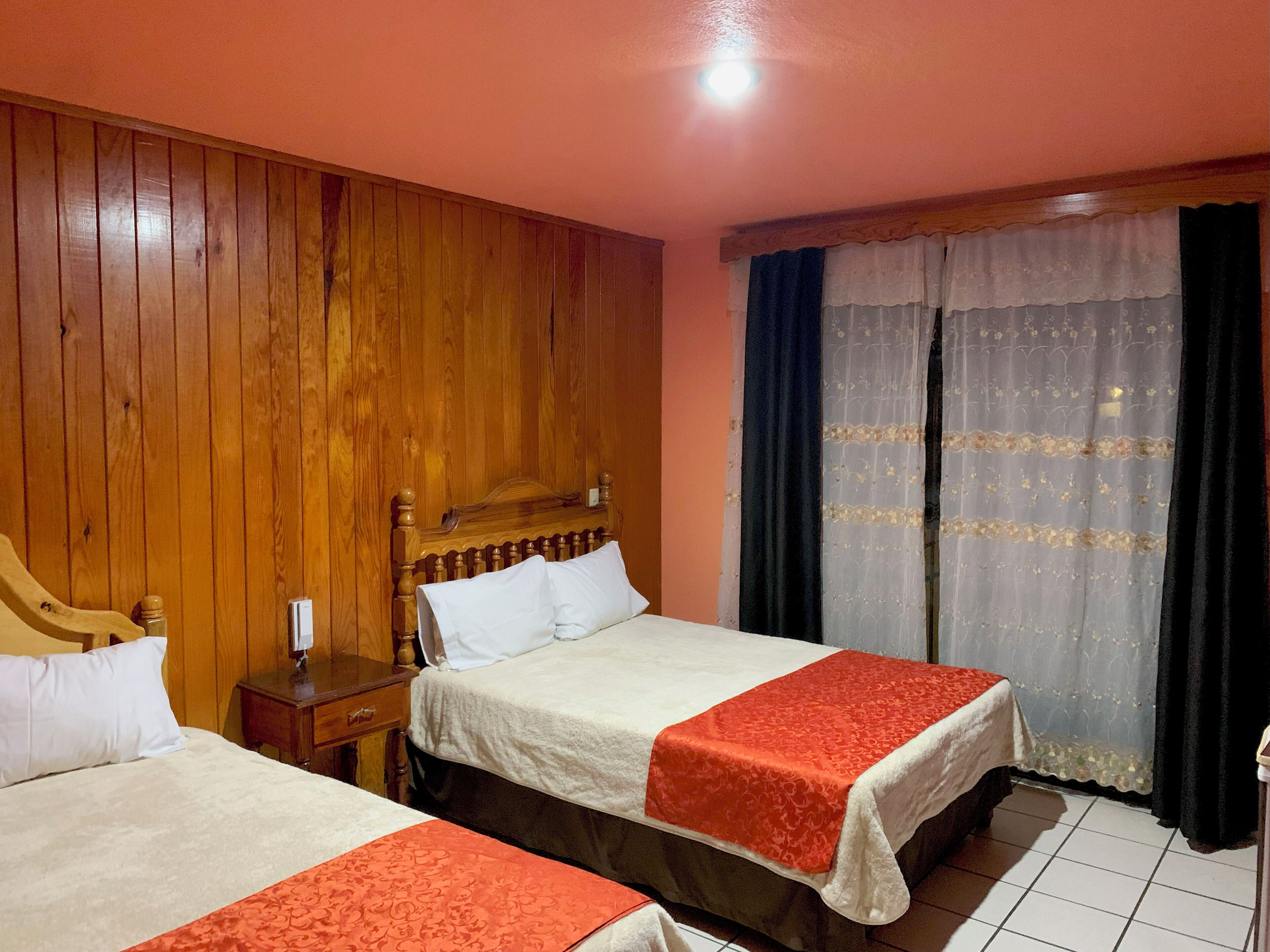 Habitación de hotel con dos camas, paneles de madera en las paredes, una mesita de noche y una ventana con cortinas.