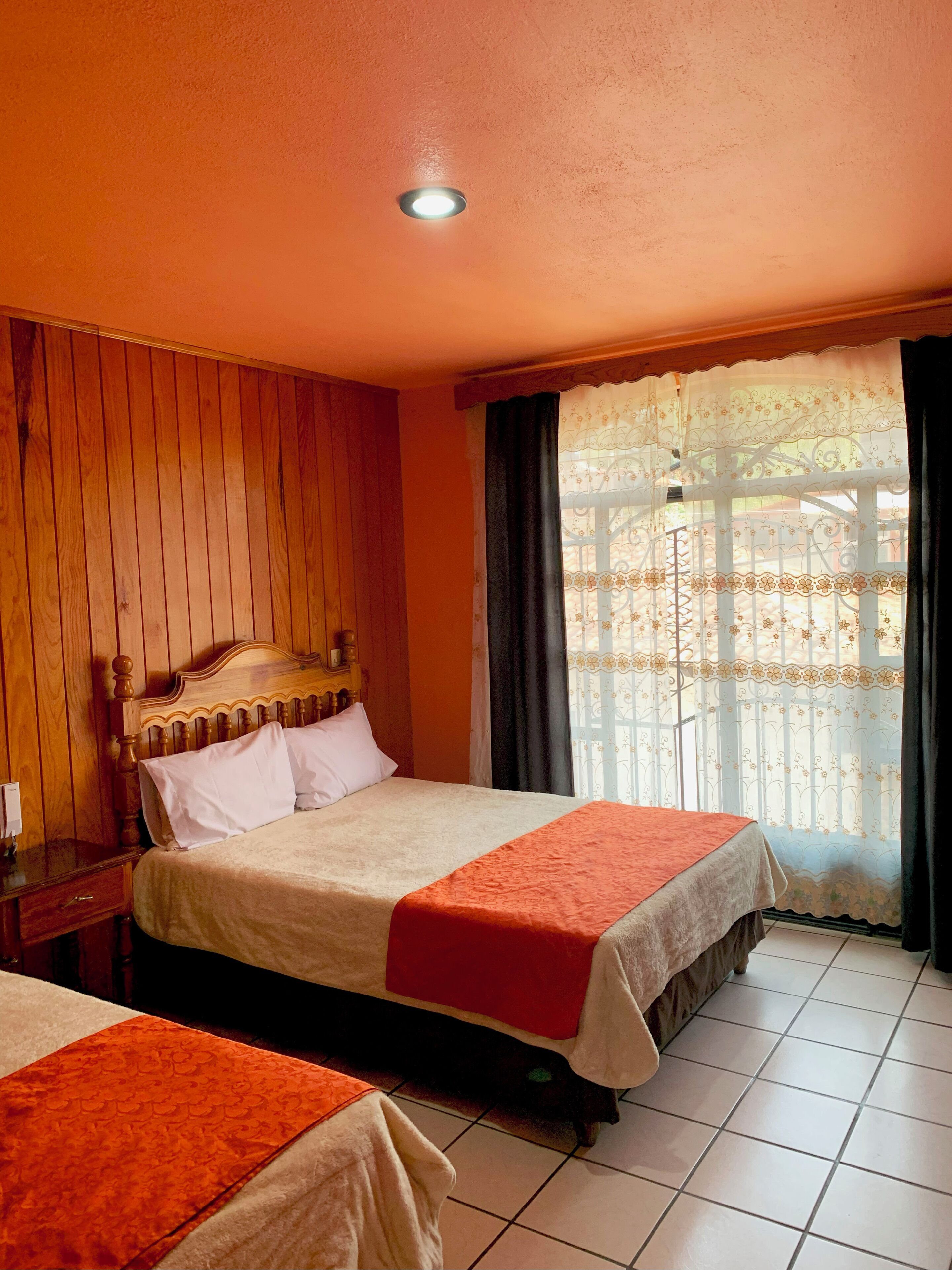 A hotel room with a wooden headboard, a bed with a white and orange bedspread, a nightstand with a lamp, and a window with a curtain.