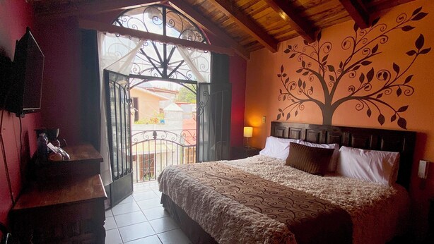 Een slaapkamer met een groot bed, een houten hoofdbord, een muurschildering, een balkon met een sierijzeren hek en een kleine tafel met een lamp.
