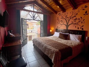Un dormitorio con una cama grande, cabecera de madera, un mural en la pared, un balcón con reja de hierro forjado y una pequeña mesa con lámpara.