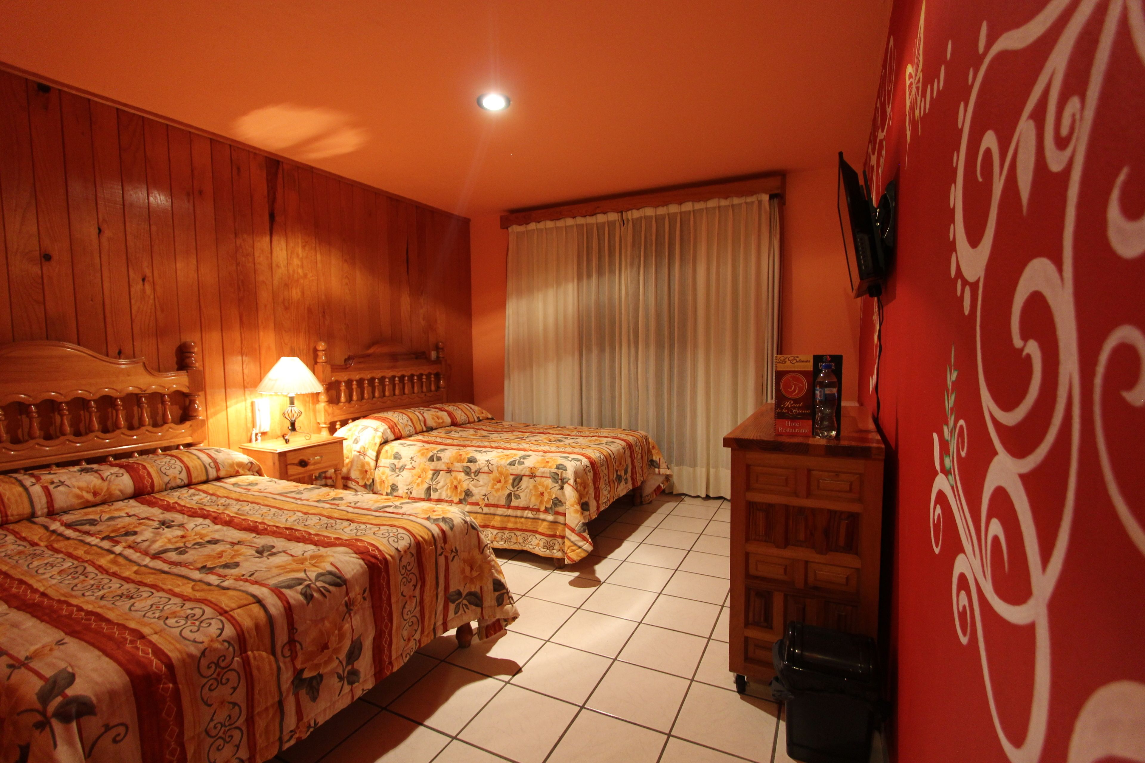 Habitación de hotel con dos camas, cabecero de madera, mesita de noche con lámpara, pared roja con un diseño decorativo y ventana con cortinas.