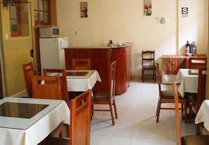 Un comedor con sillas y mesas de madera, una zona de cocina con refrigerador y microondas, y una encimera con hervidor.