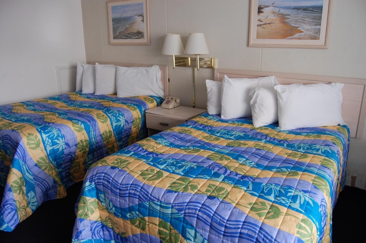 Ein Hotelzimmer mit zwei Betten, beide mit floralem Überwurf. Es gibt zwei Nachttische, einer mit einer Lampe und der andere mit einem Telefon. An den Wänden hängen gerahmte Bilder von Stränden.