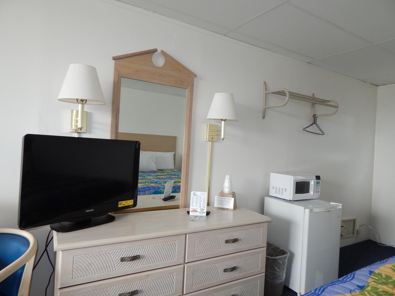 A hotel room with a bed, a desk with a computer, a television, a mirror, a lamp, a microwave, and a small trash bin.