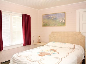 A bedroom with a bed, a nightstand, a lamp, a window with curtains, and a painting on the wall.