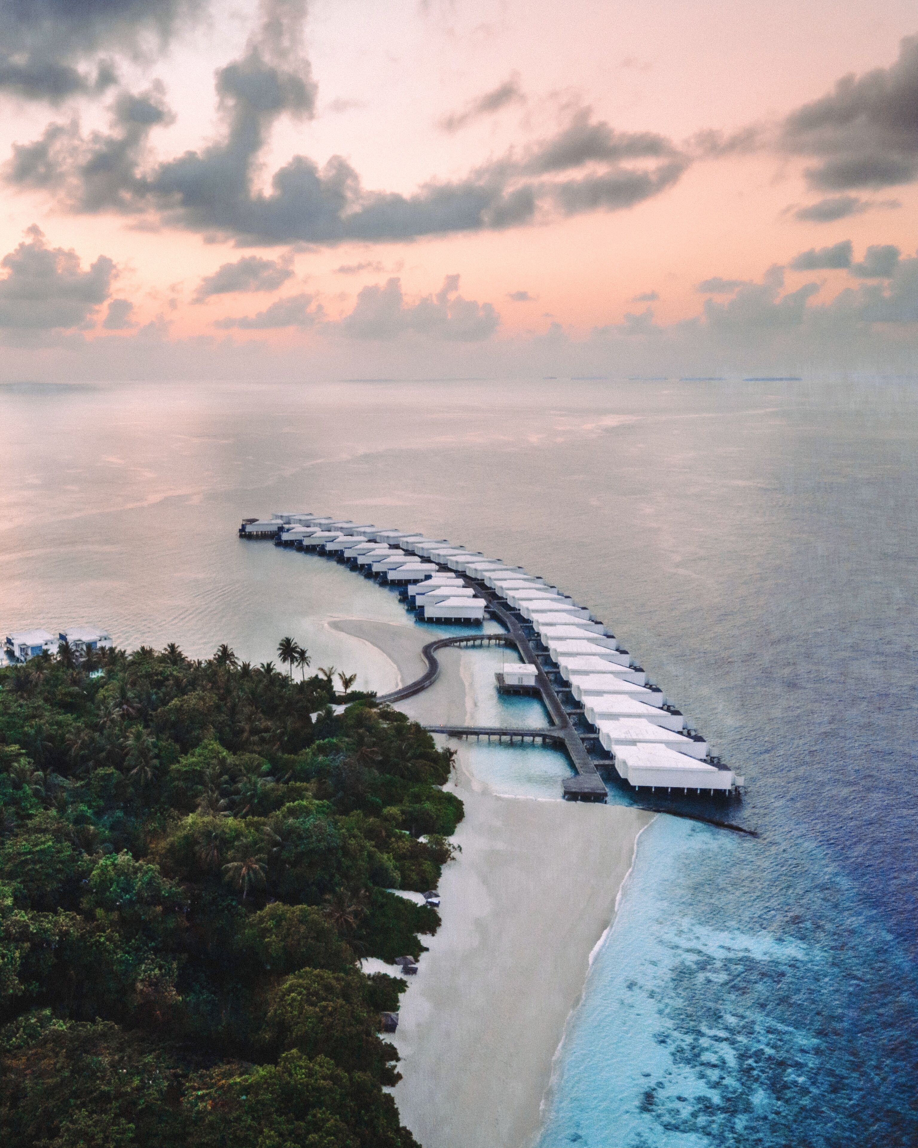 An aerial view of a long, curved overwater resort with multiple levels of white structures, connected by walkways, surrounded by a sandy beach and dense tropical vegetation.