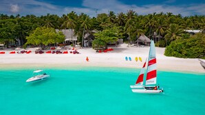 A beach with a sailboat, palm trees, and beach umbrellas. - Amilla Maldives (Finolhas Island)