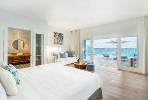 Habitación de hotel con una cama grande, un sofá, un espejo redondo, una mesita de madera y vistas al océano a través de puertas de cristal amplias.